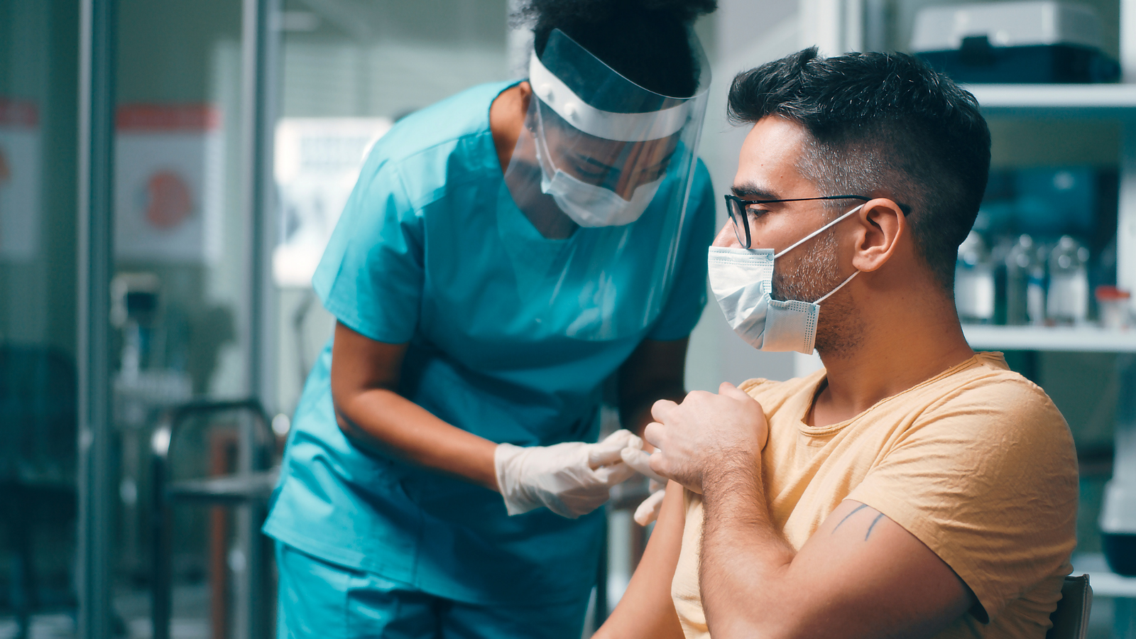 Photo of a woman in blue medical scrubs wearing a surgical mask and face shield injecting needle into the arm of a man wearing glasses and a surgical mask in a light orange t-shirt