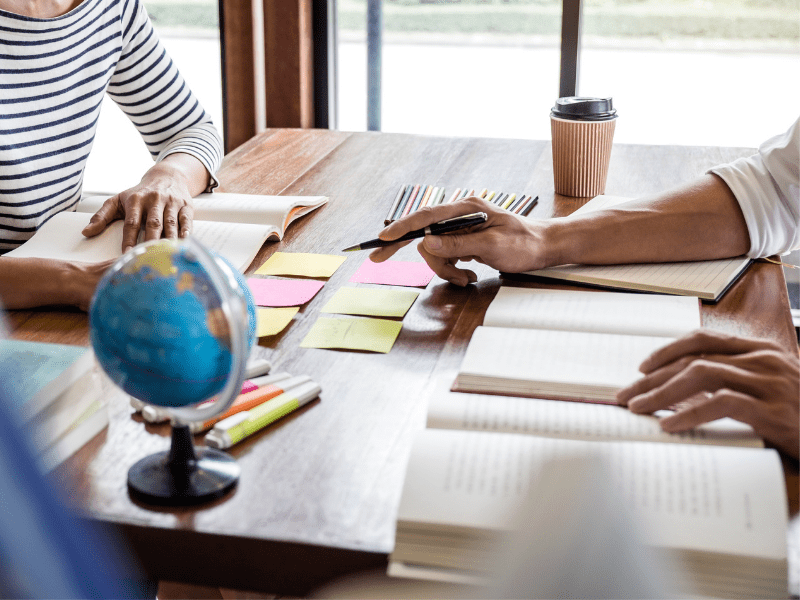 photo of two people's hands on opposite sides of a table that has books, post-it notes, and a globe on it