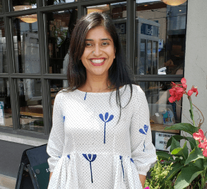 Kavita is on the sidewalk wearing a white dress posing outside a store/restaurant. Some red and purple flowers can be seen behind her. The glass windows of the store/restaurant reflect a bit of the opposite side of the street.