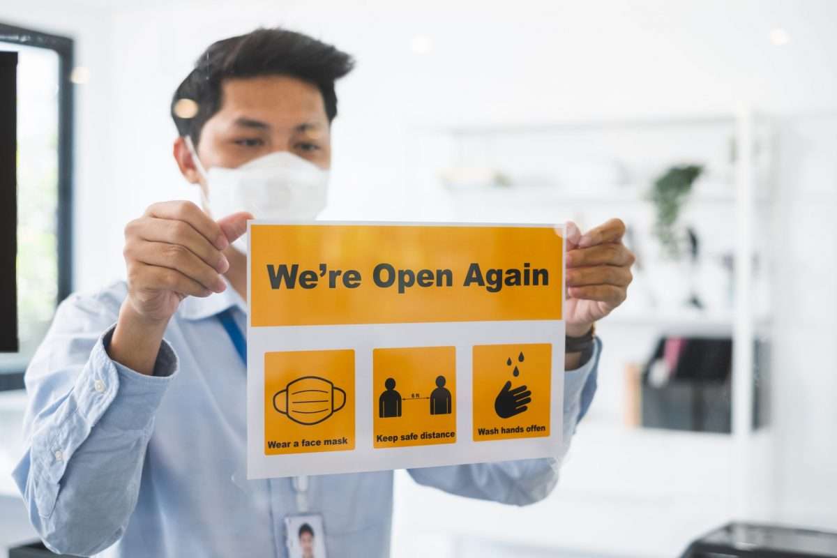 An employee in an office holding a sign that says, "We're open again," and has icons that suggest wearing a mask, watching your distance and washing your hands.